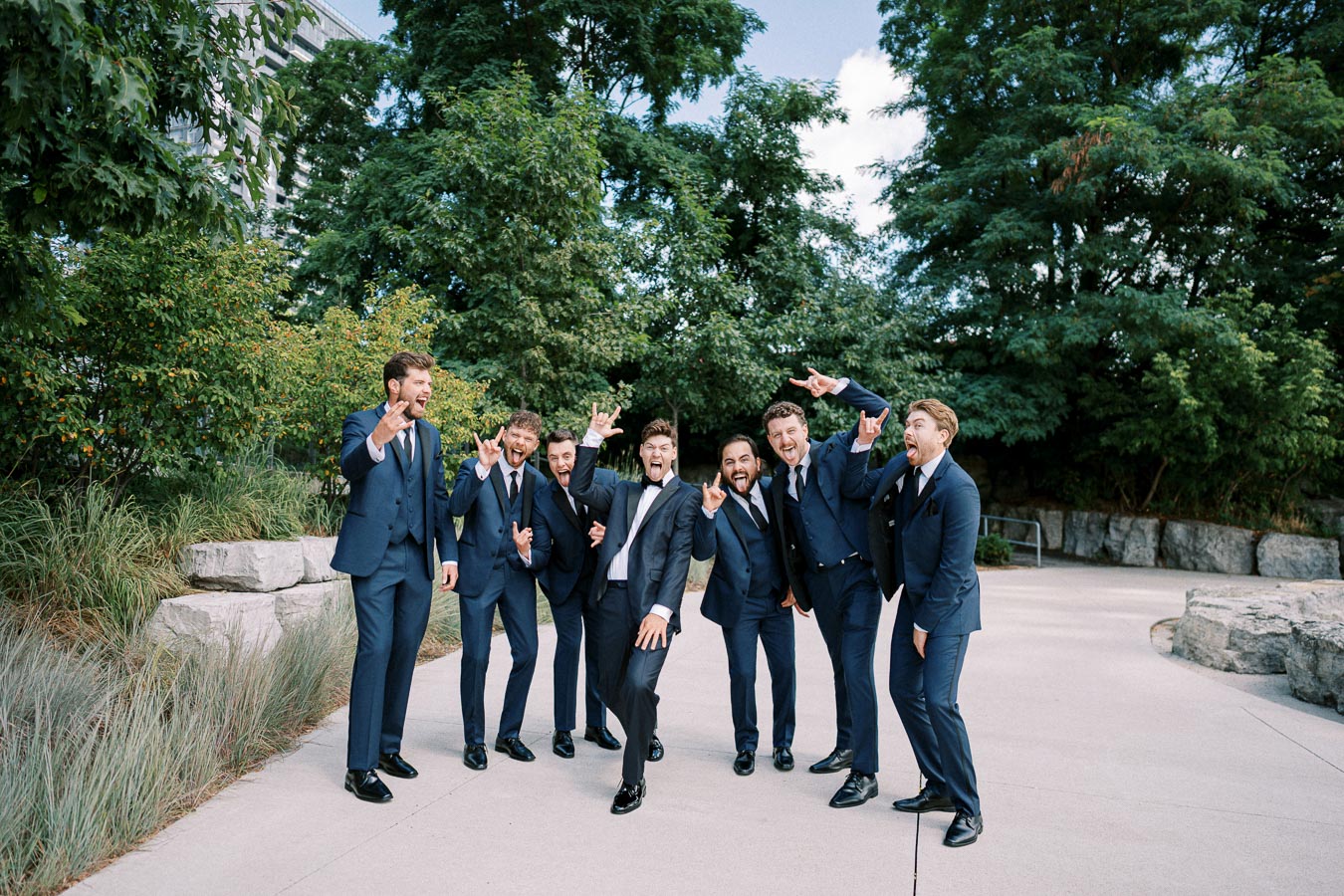 A group of groomsmen in matching blue suits posing playfully with enthusiastic expressions and hand gestures, standing outdoors on a paved path surrounded by lush greenery and large rocks.