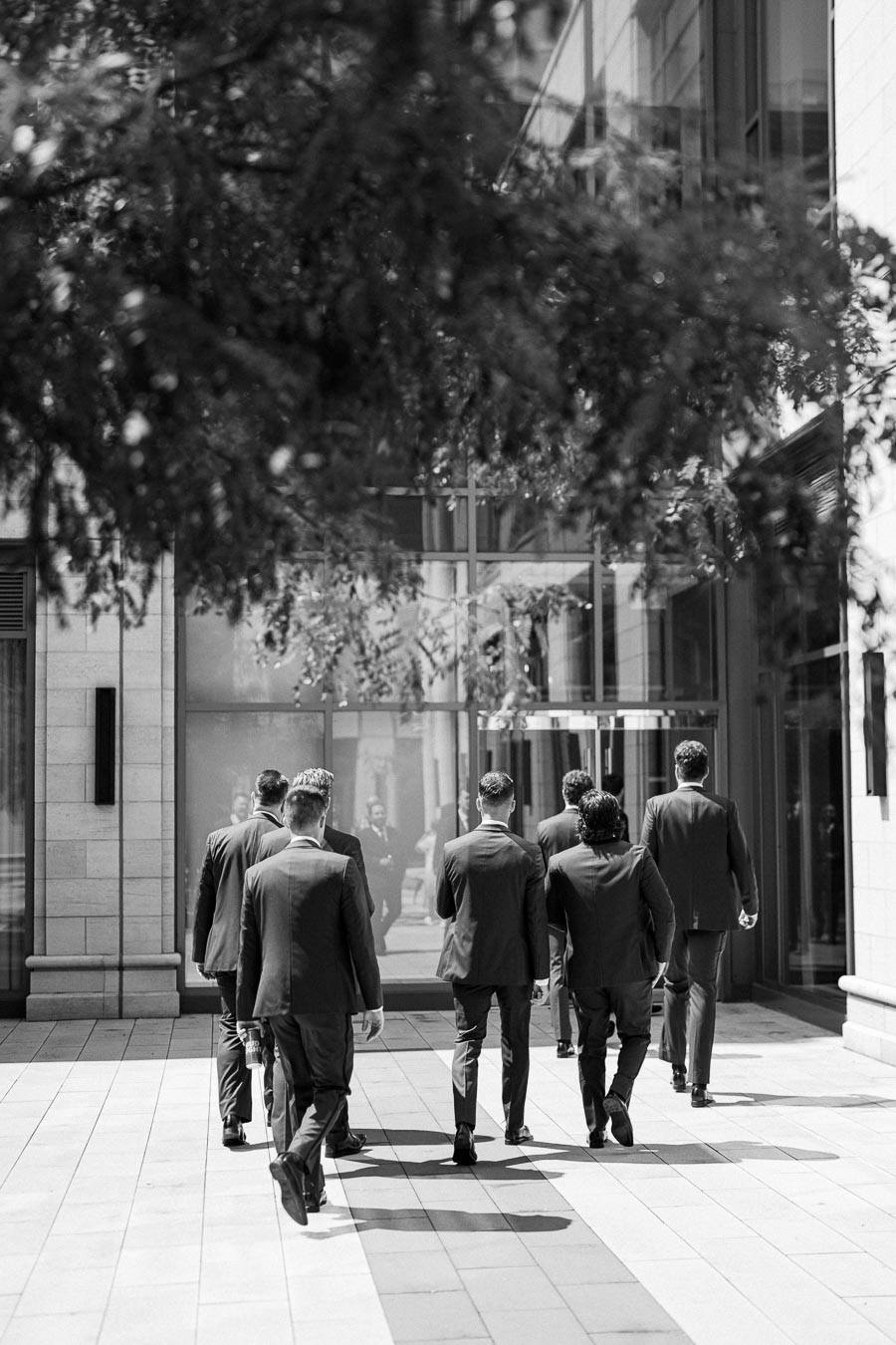 A group of men in suits walking towards a modern building entrance, surrounded by urban architecture and leafy trees, in black and white.