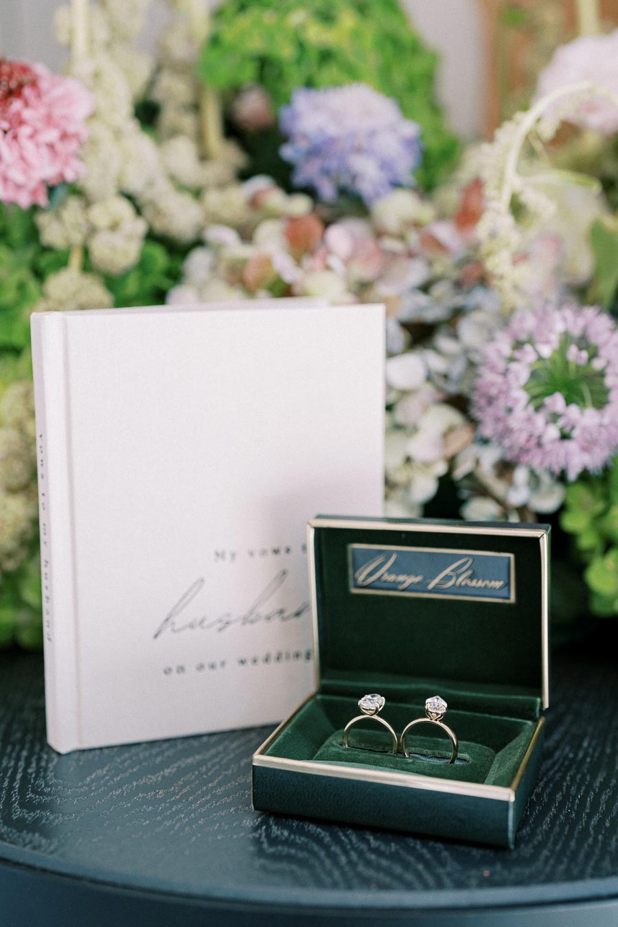 Elegant wedding vow book and diamond rings in an Orange Blossom box placed on a table with a floral background.