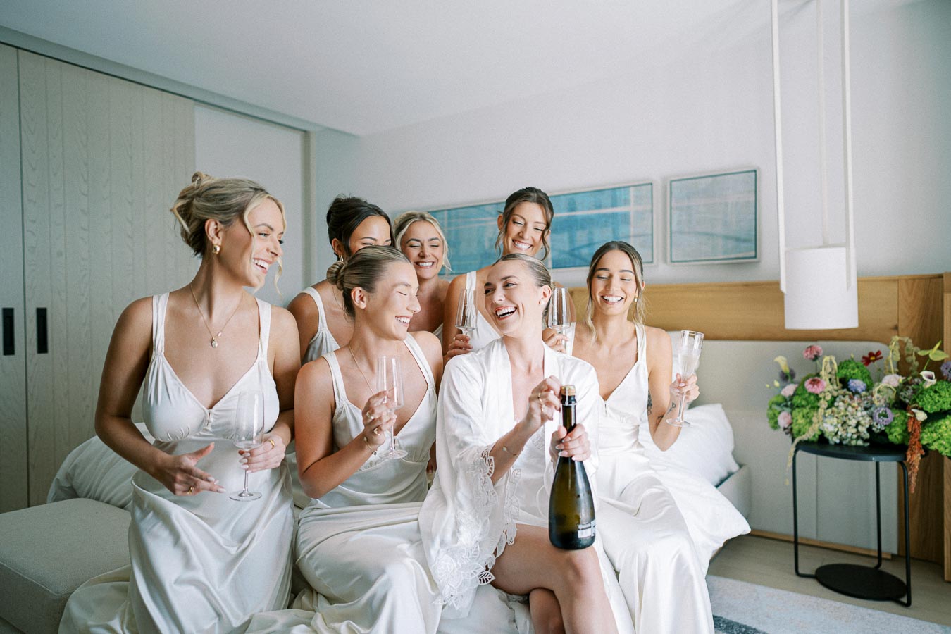 A joyful bridal party of women in matching satin gowns sharing a celebratory moment with champagne on a wedding morning in a stylish, modern bedroom setting.
