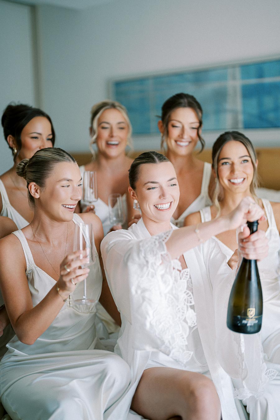 Bridesmaids and bride in white robes celebrating with champagne in a bright room, smiling and enjoying a pre-wedding gathering.