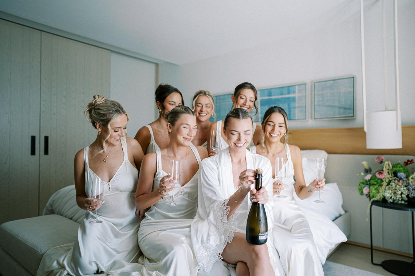 Bridesmaids in matching robes and dresses celebrate with champagne in a modern hotel room, creating joyful pre-wedding memories.