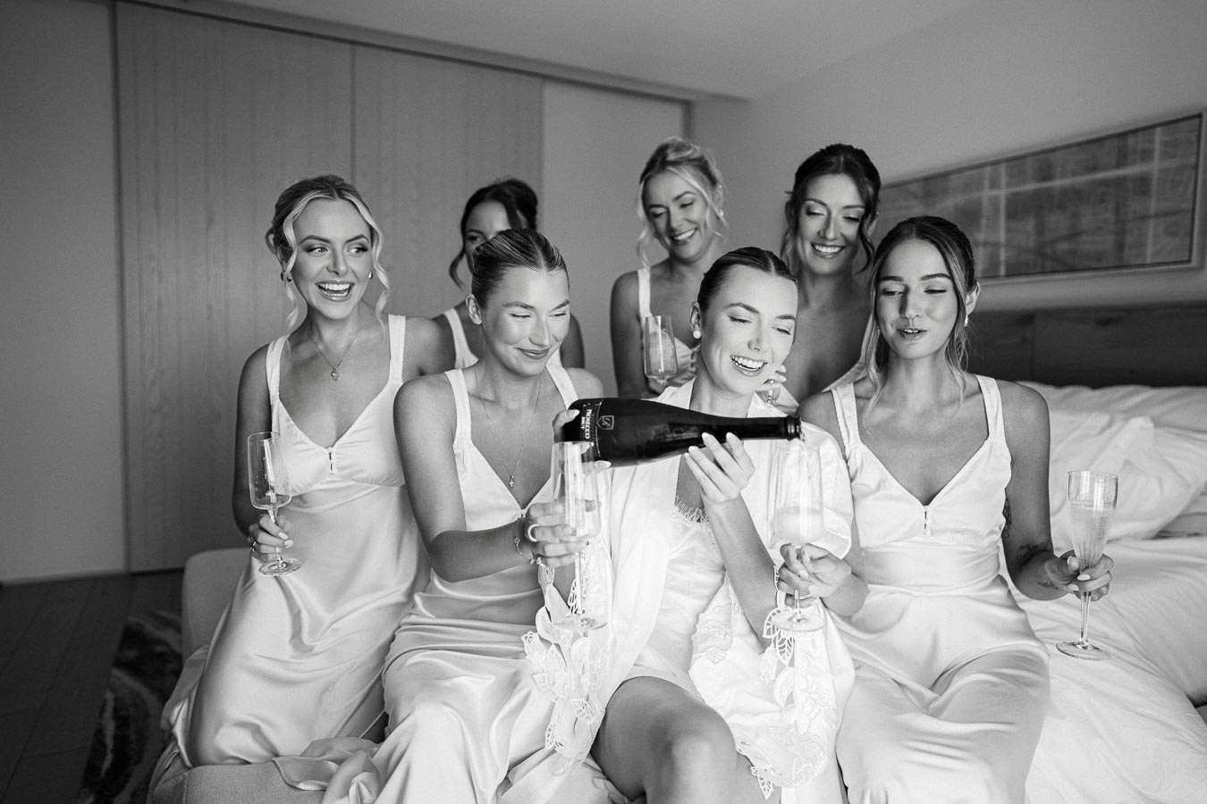 Bridesmaids in matching dresses celebrating with champagne in a bridal suite, laughing and smiling, before a wedding ceremony.