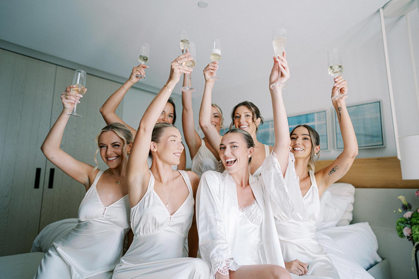 Group of smiling bridesmaids in white robes joyfully raise champagne glasses in a modern, light-filled bedroom setting, celebrating a special occasion.