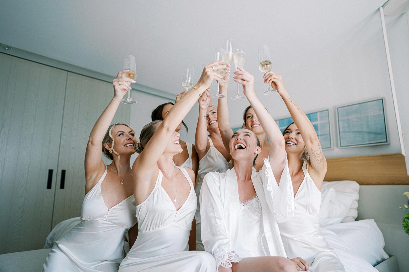 Bridesmaids in white robes raising champagne glasses, celebrating in a modern bedroom setting.