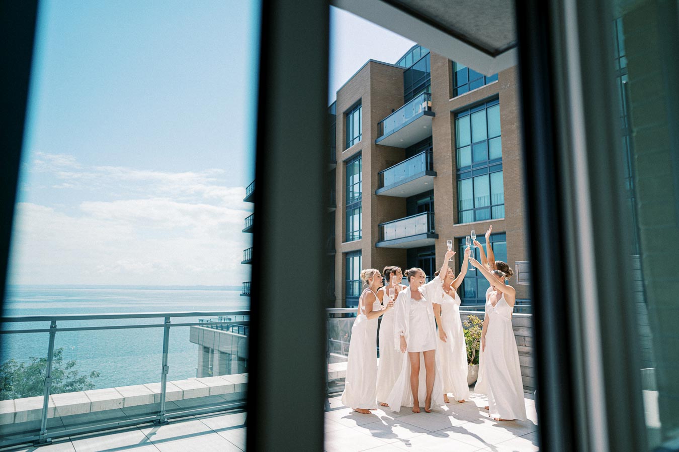 A group of women celebrating on a modern balcony overlooking the ocean, with high-rise buildings in the background and a clear blue sky.