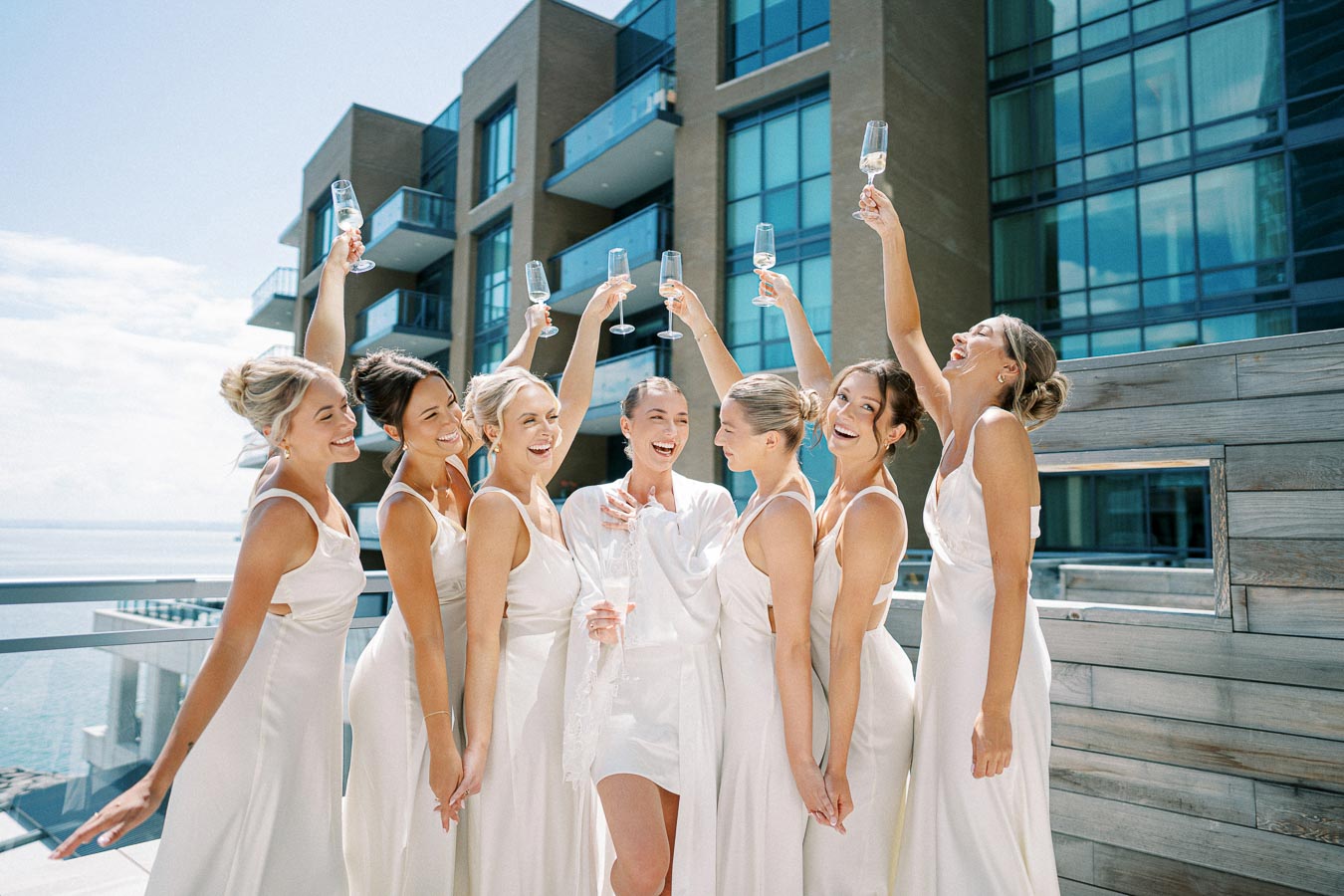Bridesmaids in elegant white dresses joyfully raising champagne glasses on a sunny rooftop terrace, with modern buildings and ocean view in the background.