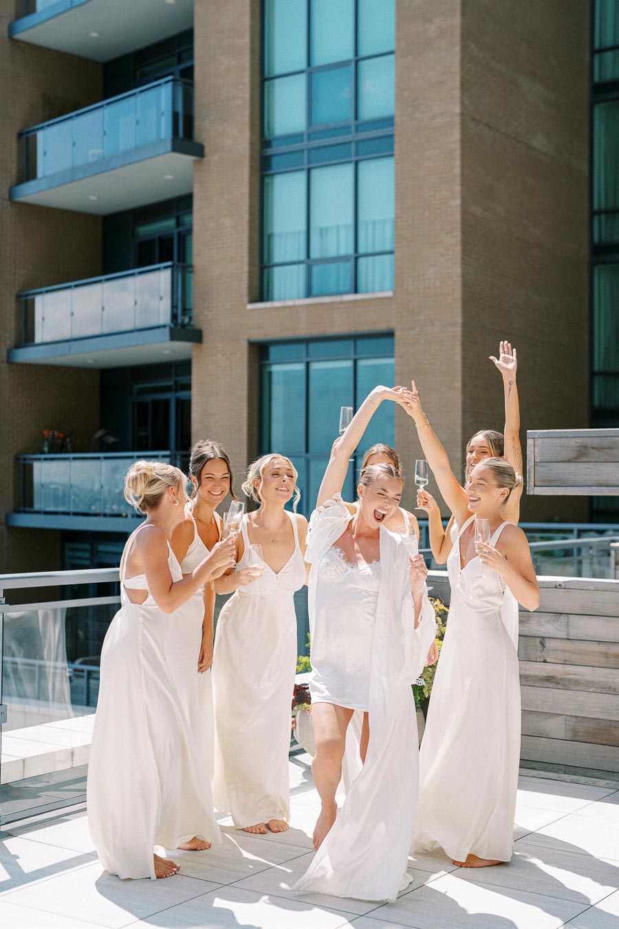 Bridal party celebration on a sunny balcony, women in elegant white dresses joyfully cheering with champagne glasses.