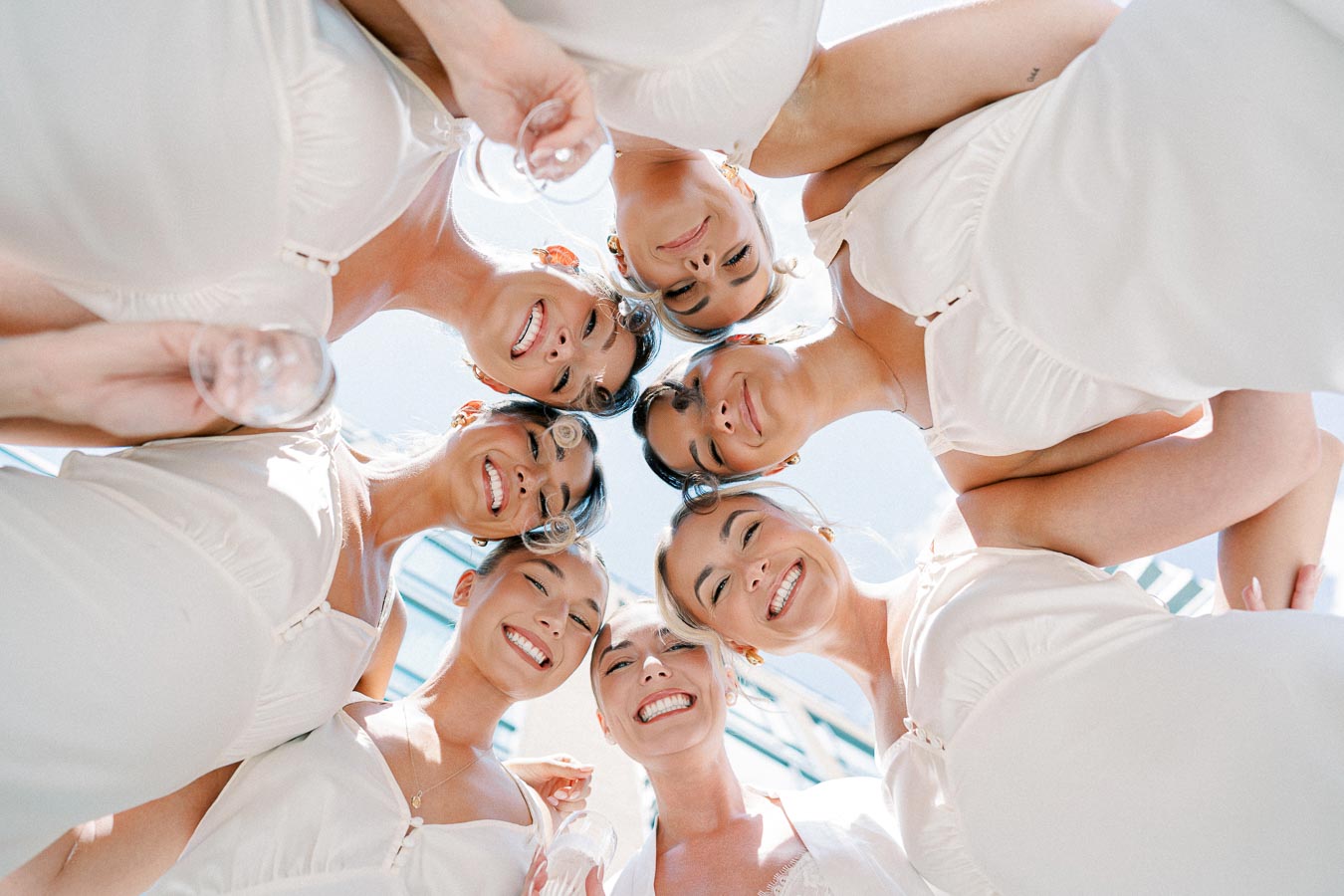 Seven smiling bridesmaids in white dresses captured from a low-angle perspective, celebrating outdoors under a clear blue sky.