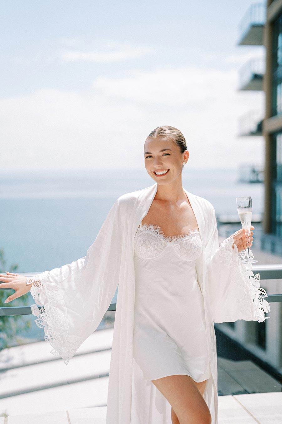 Smiling woman in white lace gown holding a glass of champagne on a sunny balcony with ocean view.