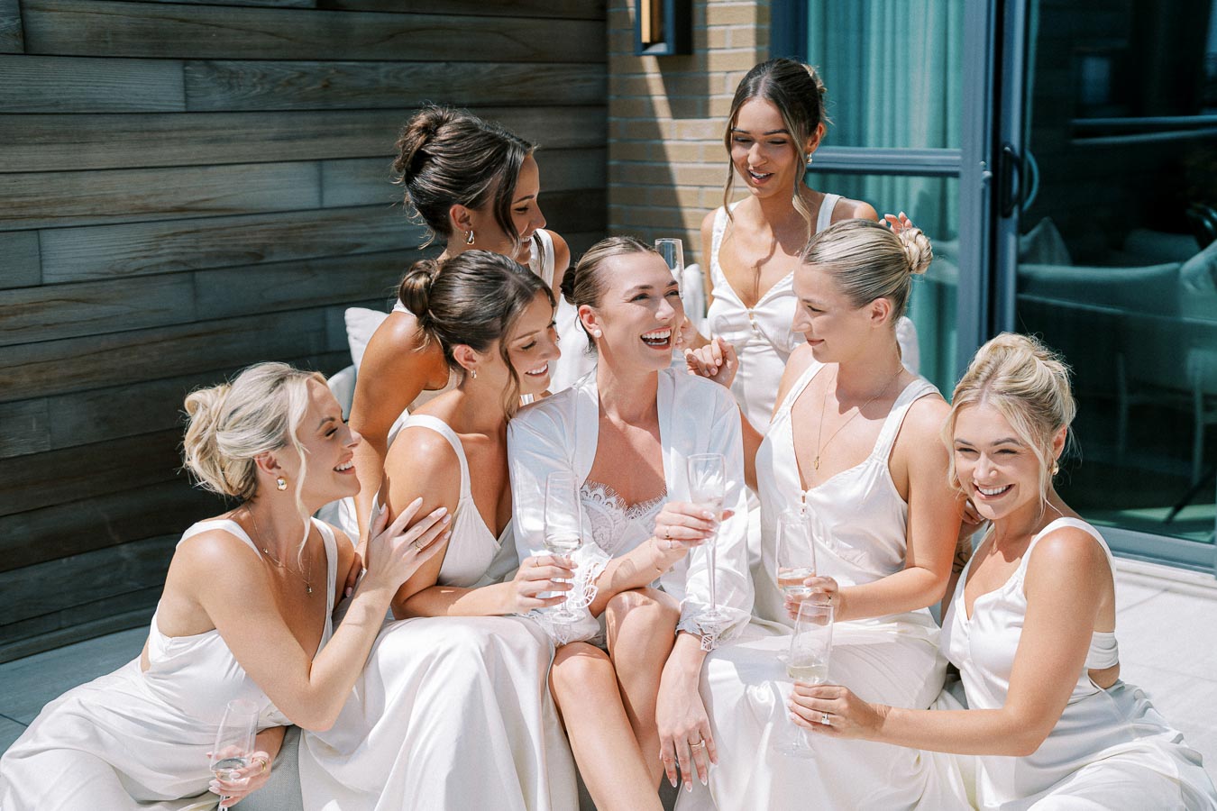 Elegant bridal party laughing and enjoying champagne on a sunny day, wearing white dresses on a modern patio.
