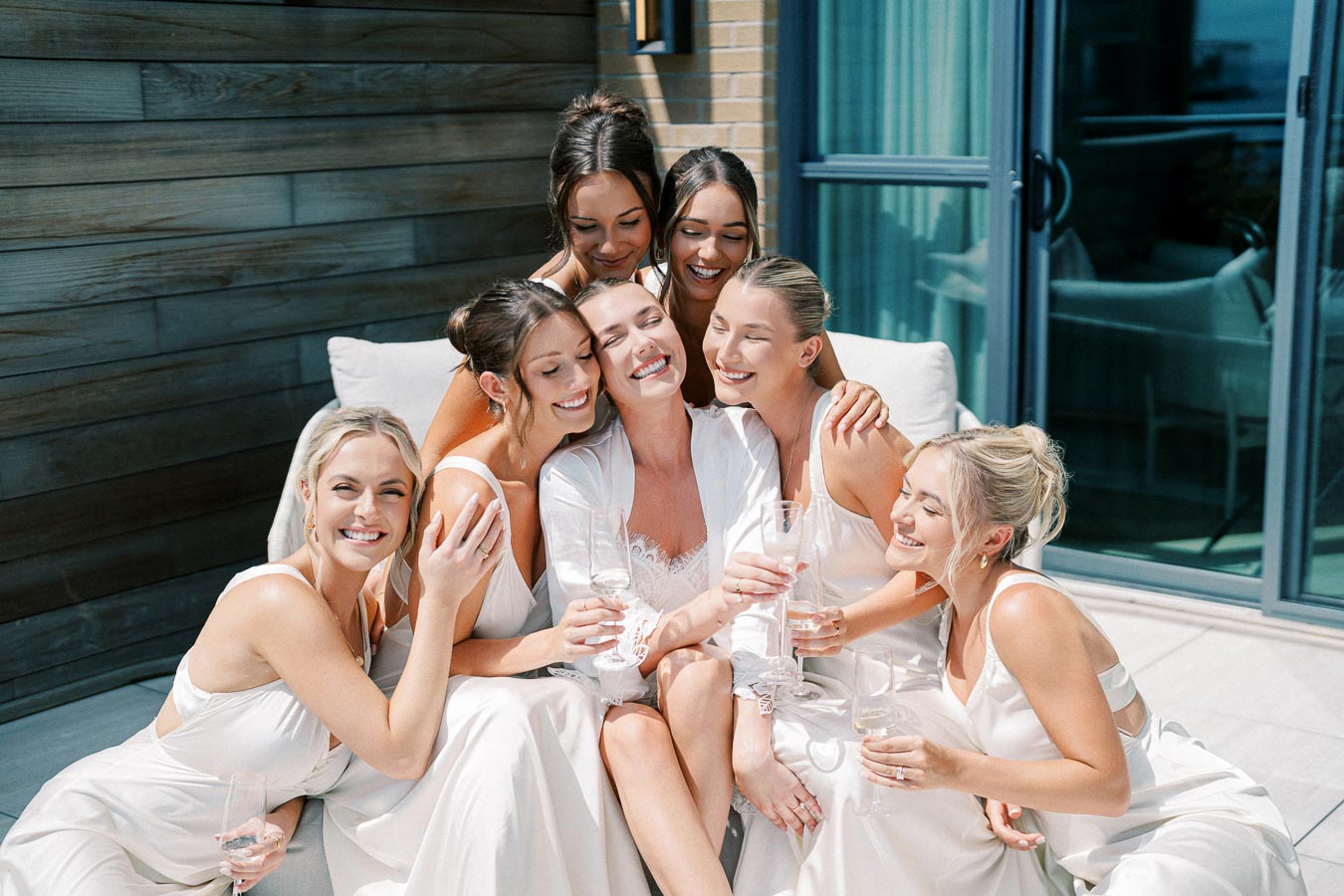 A group of smiling bridesmaids in elegant white dresses, sitting together with champagne glasses on a sunny patio.