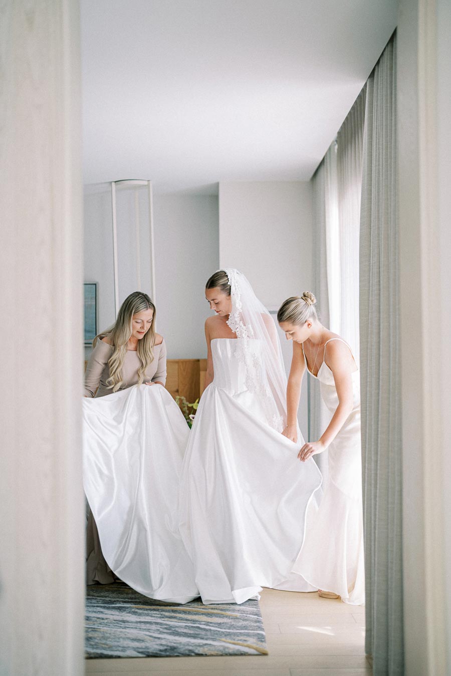 Three women, including a bride in a strapless white wedding gown with a lace veil, gracefully preparing for a wedding ceremony in a bright, softly lit room.