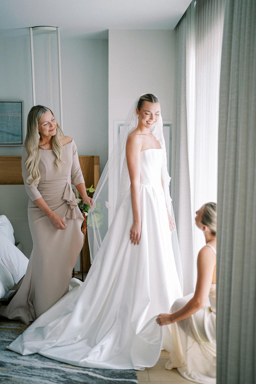Bride in elegant white wedding gown and veil stands near window, smiling, with two bridesmaids in neutral dresses assisting.