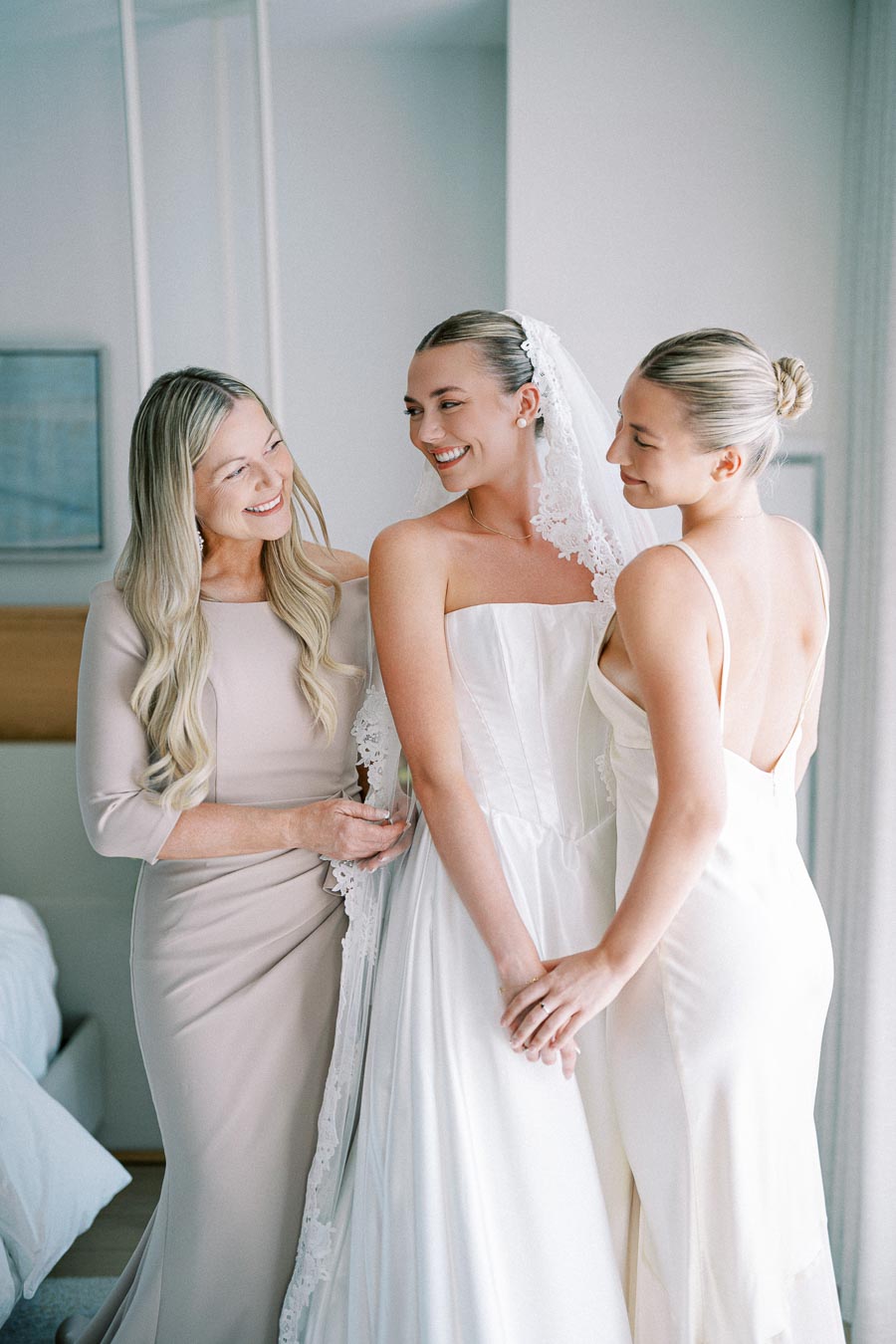 Bride wearing a white wedding dress shares a joyful moment with two women, both in elegant dresses, in a bright room.