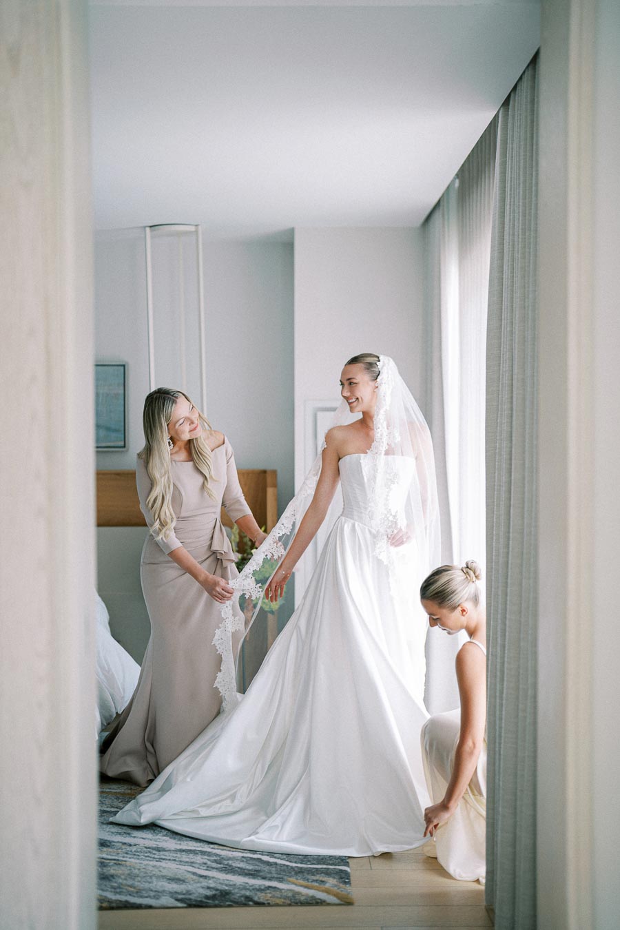 Bride getting ready for her wedding with the help of two bridesmaids, one adjusting her veil and the other arranging the gown, in a softly lit room.