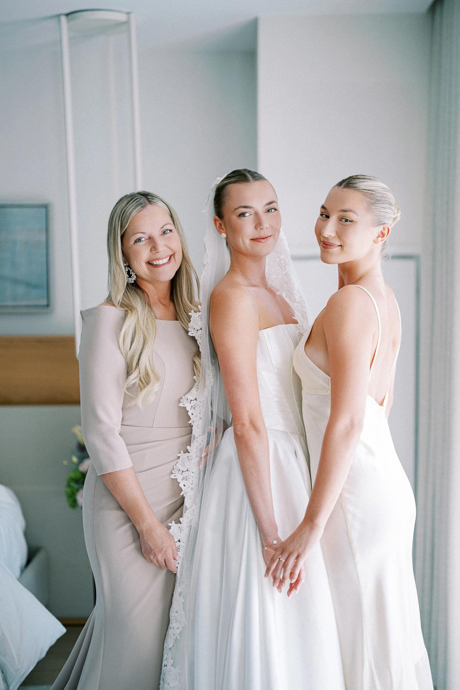 Three women smiling and posing together, with one wearing a bridal gown and veil, suggesting a wedding preparation scene indoors.