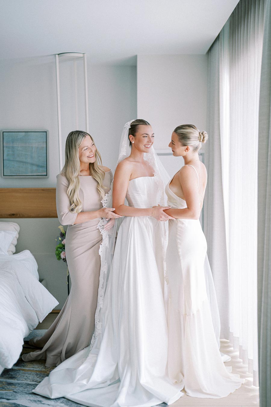 Three women in elegant gowns stand inside a softly lit room. One wears a bridal gown and veil, while the others wear long dresses. They are smiling and standing near a window, sharing a joyful moment before a wedding ceremony.