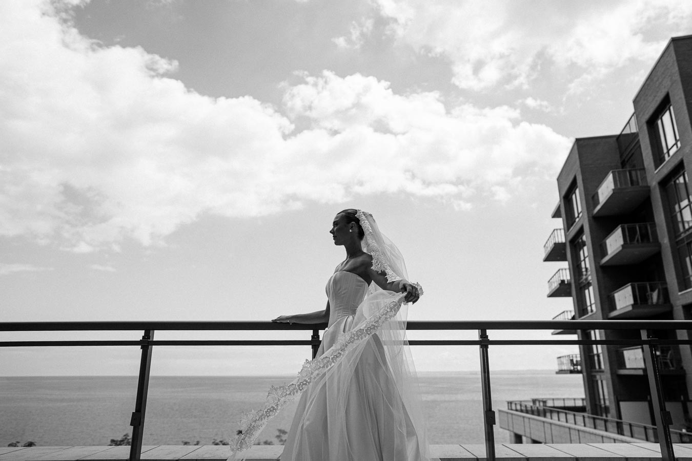 Black and white image of a bride in a flowing gown and veil standing on a balcony, overlooking the ocean with modern buildings in the background and a cloudy sky above.