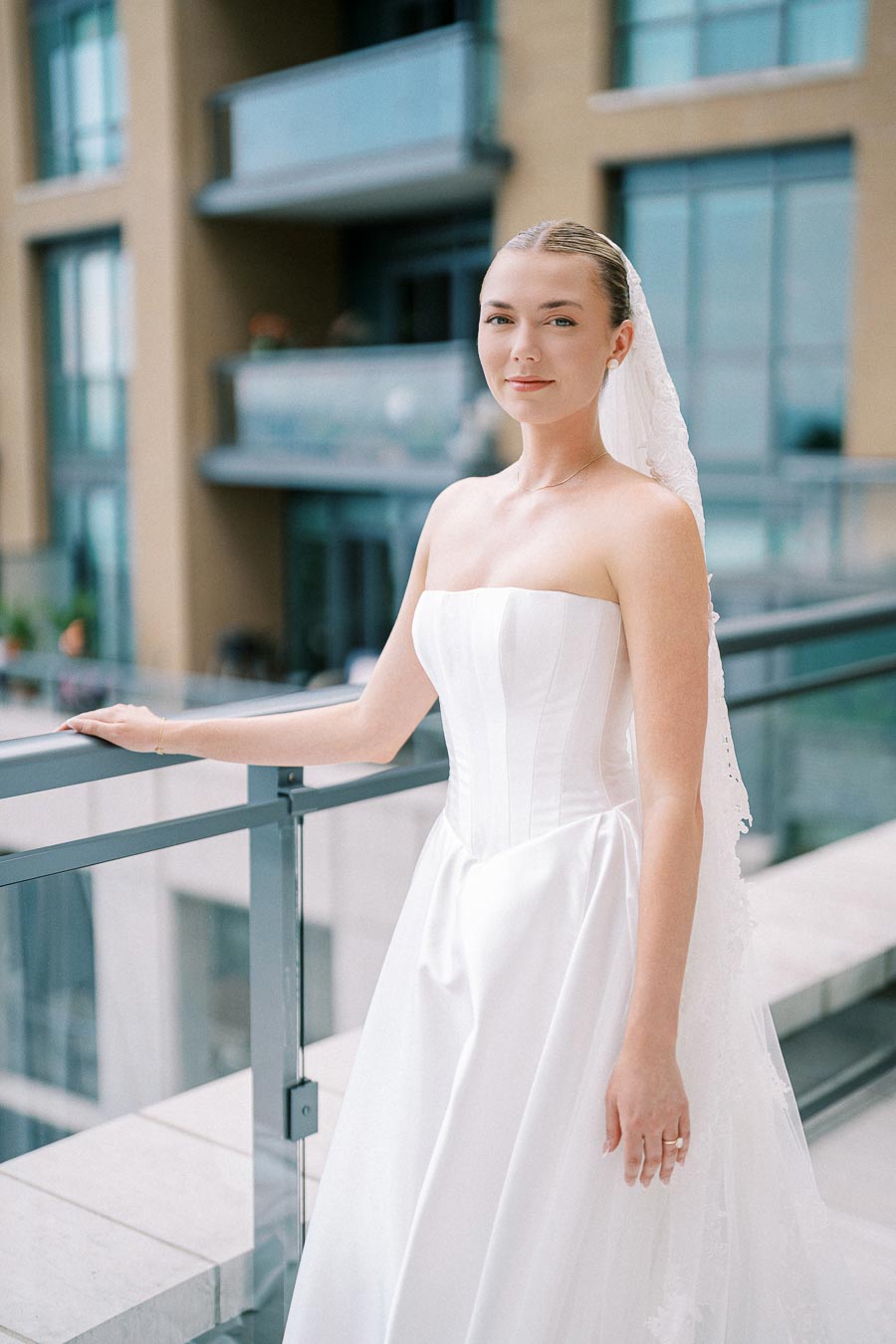 Young bride in elegant white wedding gown standing on a terrace with modern building in the background.