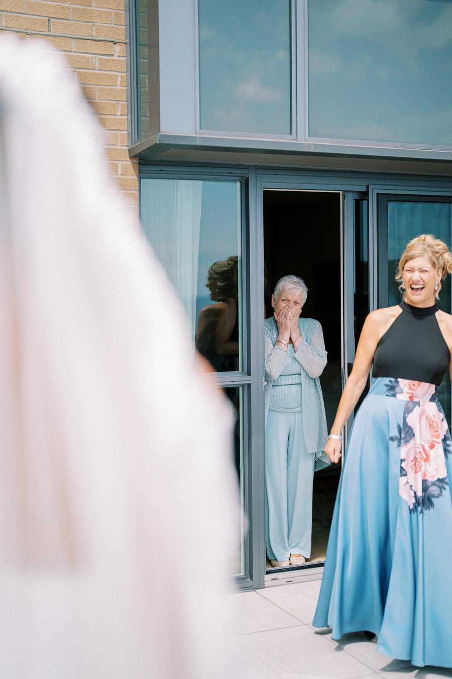 Two women standing in the doorway of a building, one with a joyful expression wearing a floral skirt, while the other, an elderly woman, looks surprised with her hands over her mouth. The view hints at a joyful occasion, possibly a wedding or celebration.