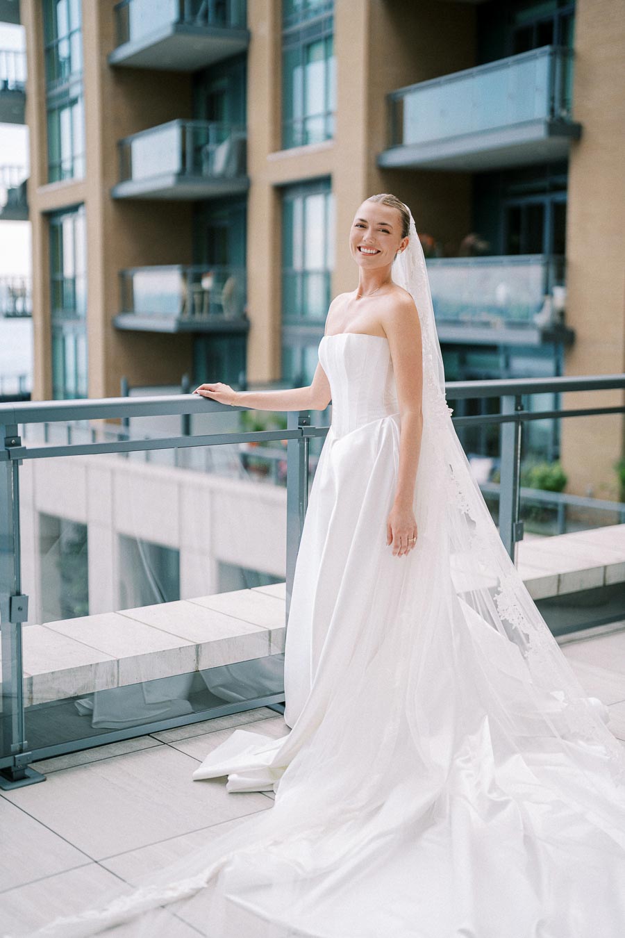 Smiling bride in elegant white wedding dress with long veil poses on modern hotel balcony with cityscape background.