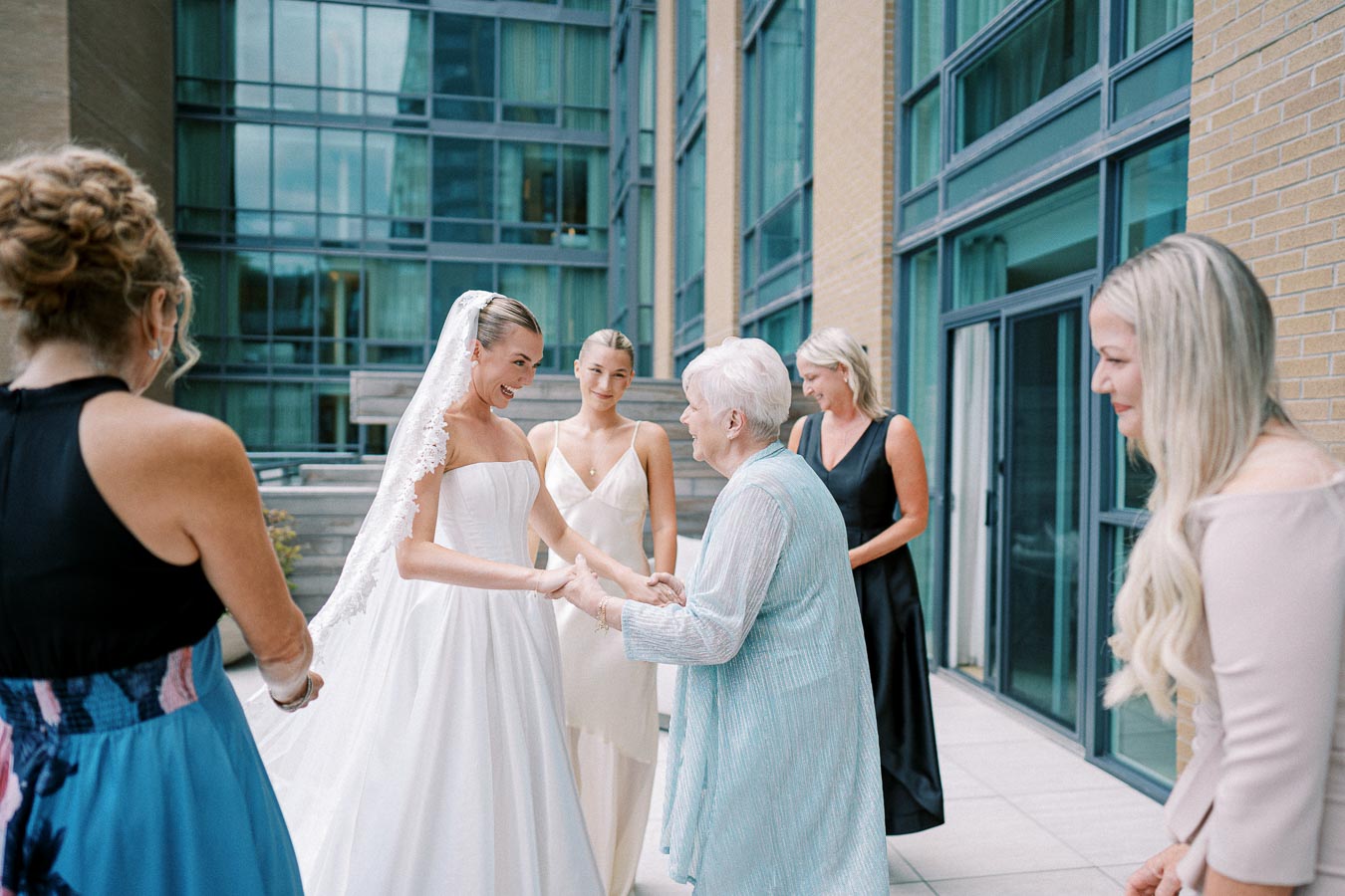 A bride in a white dress and lace veil smiles warmly while holding hands with an elderly woman in a light blue outfit on a modern terrace, surrounded by a group of women in formal dresses, captured moments before her wedding ceremony.