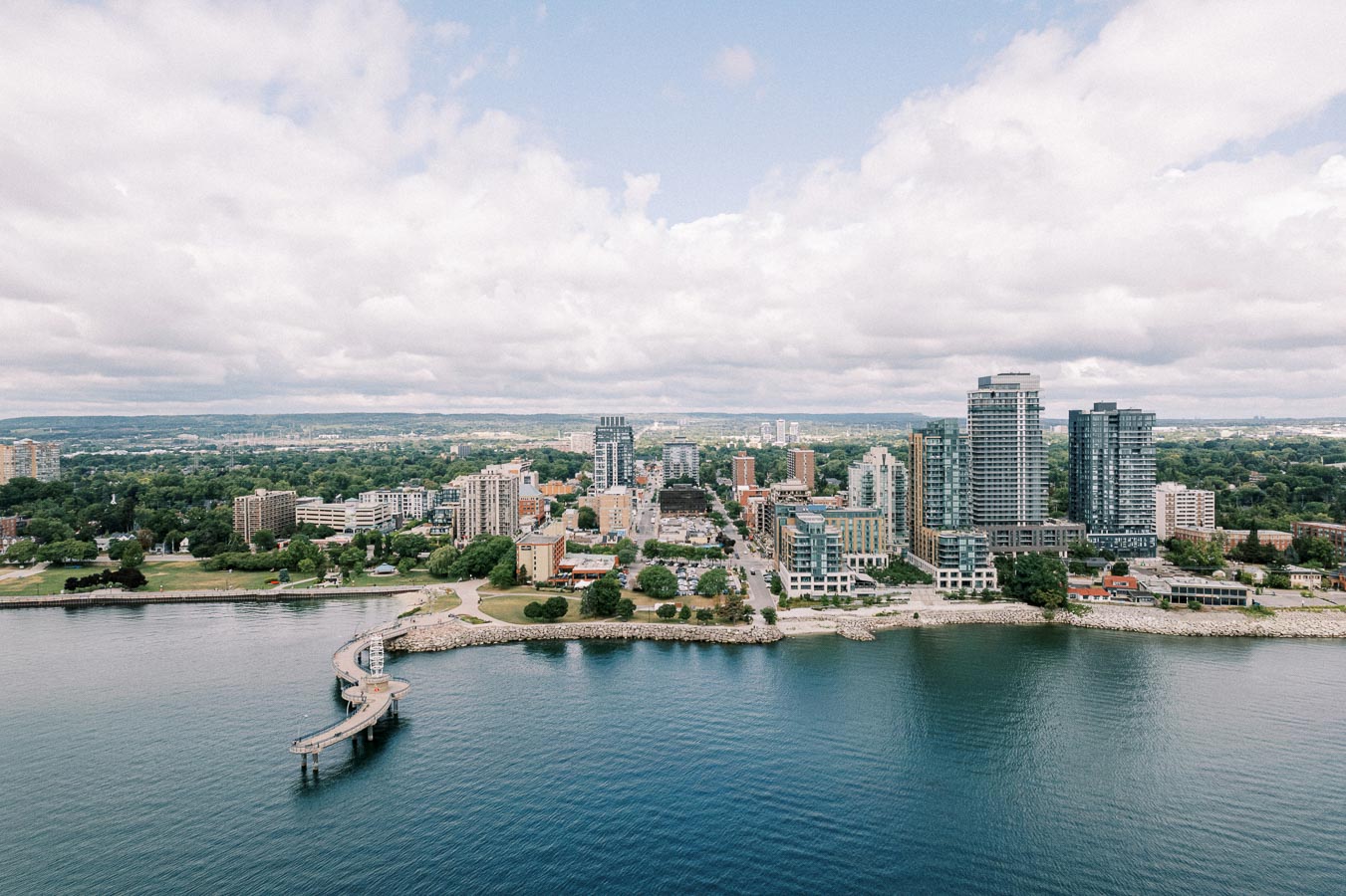 Aerial view of a coastal cityscape featuring modern high-rise buildings, lush greenery, and a curved pier extending into calm waters under a partly cloudy sky.