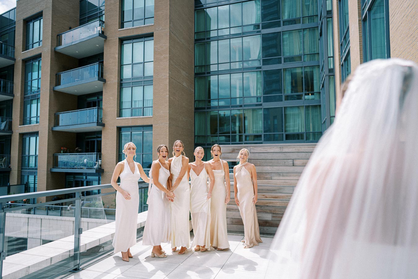 Bridesmaids in elegant blush dresses react joyfully to bride's arrival on rooftop terrace, with urban building backdrop.