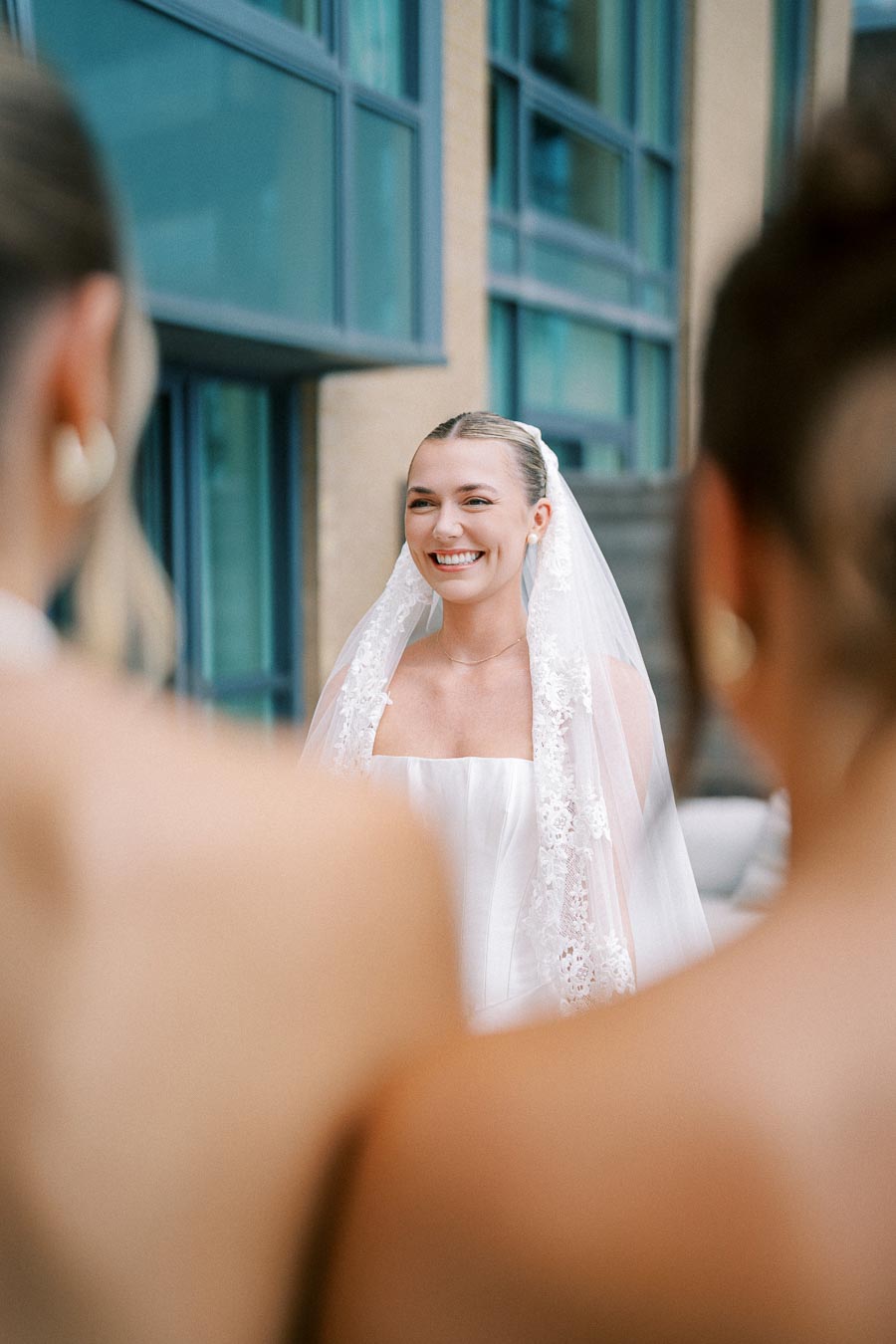 A smiling bride in a white wedding dress and lace veil stands in front of modern building windows, framed by blurred bridesmaids in the foreground.
