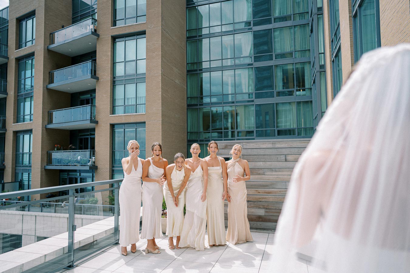 Bridesmaids in elegant white dresses react joyfully on a modern terrace as the bride approaches, with urban architecture in the background.