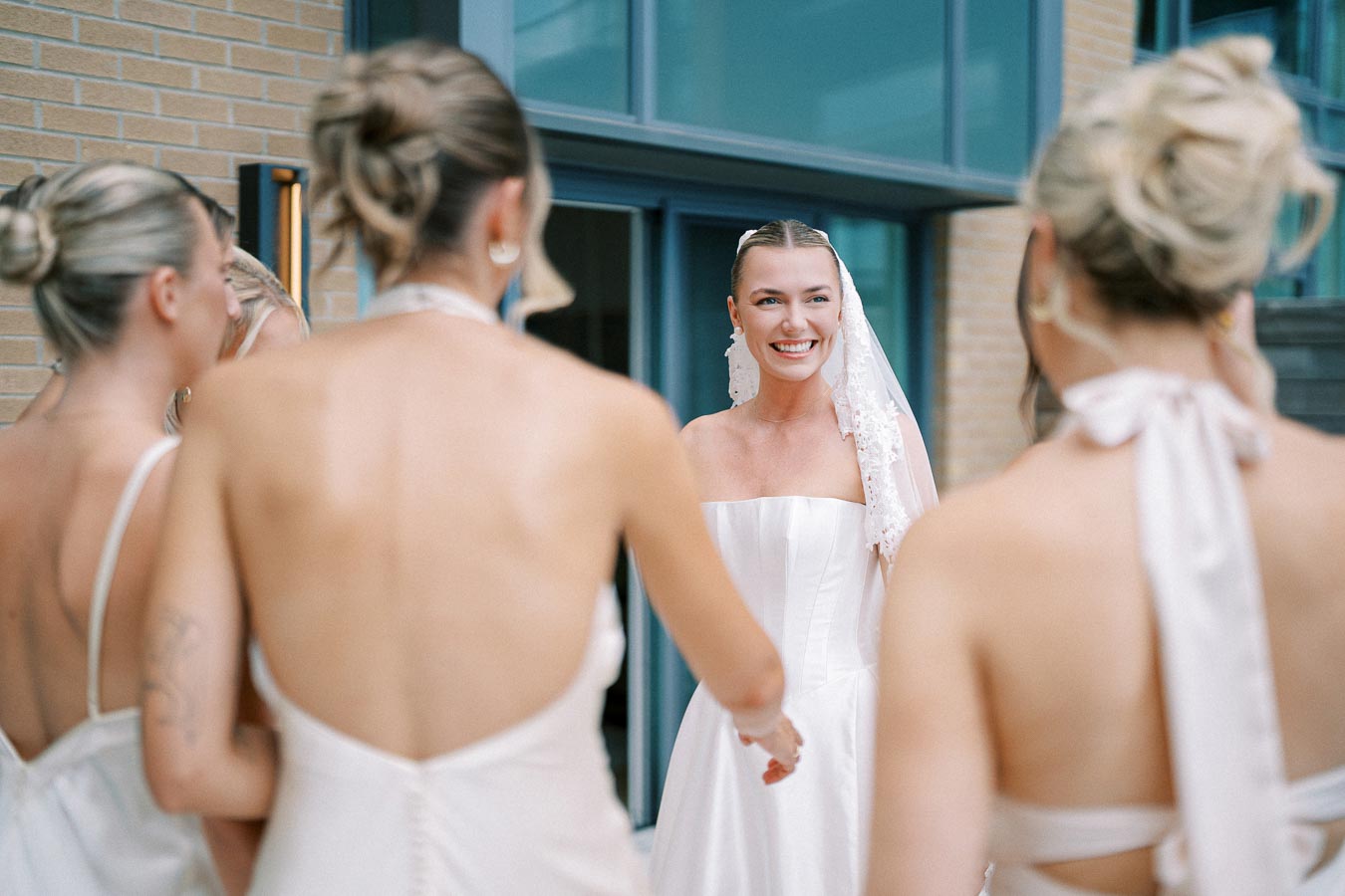 A smiling bride in a strapless white wedding dress and veil surrounded by bridesmaids in light dresses, standing outside near modern building.