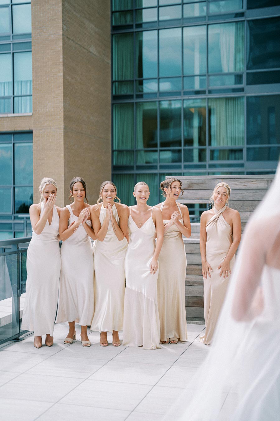 Bridesmaids in elegant matching cream dresses excitedly reacting on a modern rooftop with glass and brick architecture.
