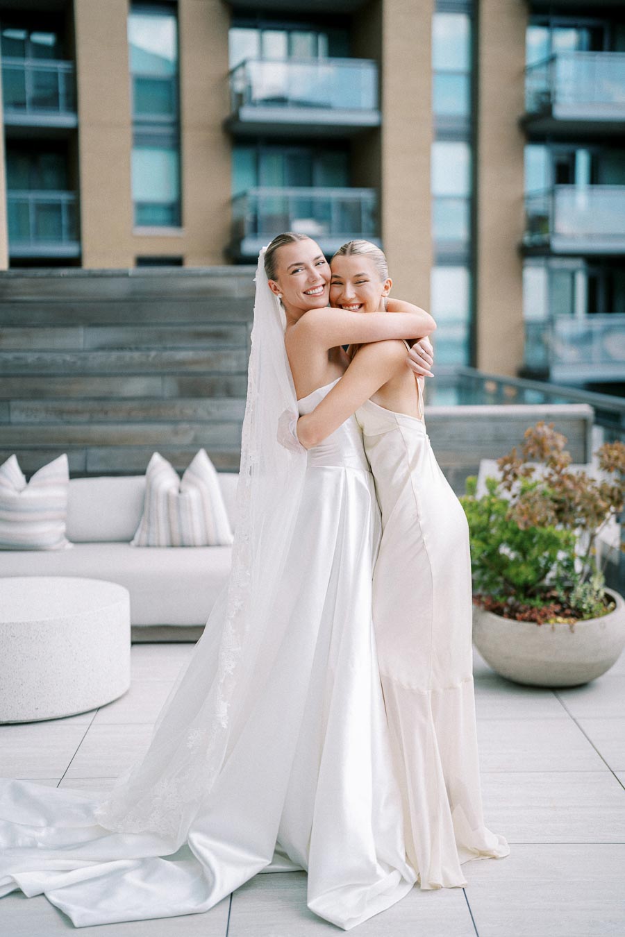 Two brides in elegant white dresses embrace joyfully on a modern patio, with apartments in the background, highlighting love and happiness on their wedding day.