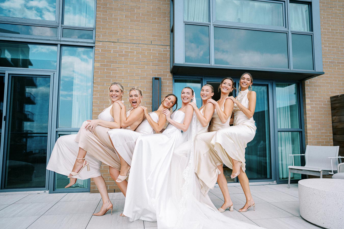 Joyful bride and bridesmaids in elegant dresses celebrate on a modern rooftop with glass and brick backdrop, wearing matching high heels.