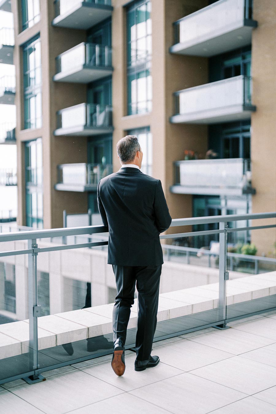 A man in a suit standing on a modern balcony, overlooking an urban residential building with glass railings and multi-level apartments.