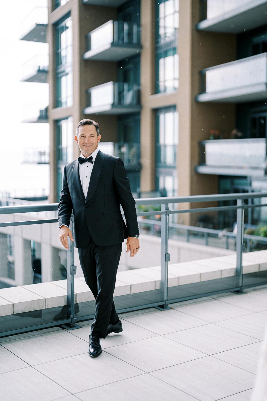 Man in a black tuxedo smiling while walking on a balcony with modern buildings in the background.