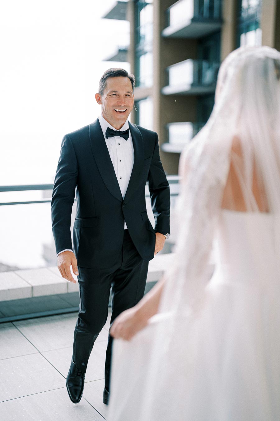 Smiling groom in black tuxedo and bow tie meeting bride in white wedding dress and veil on a modern outdoor terrace