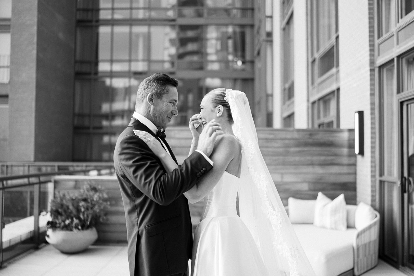 Black and white image of a joyful couple embracing on their wedding day, with the groom in a tuxedo and the bride wearing a wedding gown and veil, standing on a modern building terrace.
