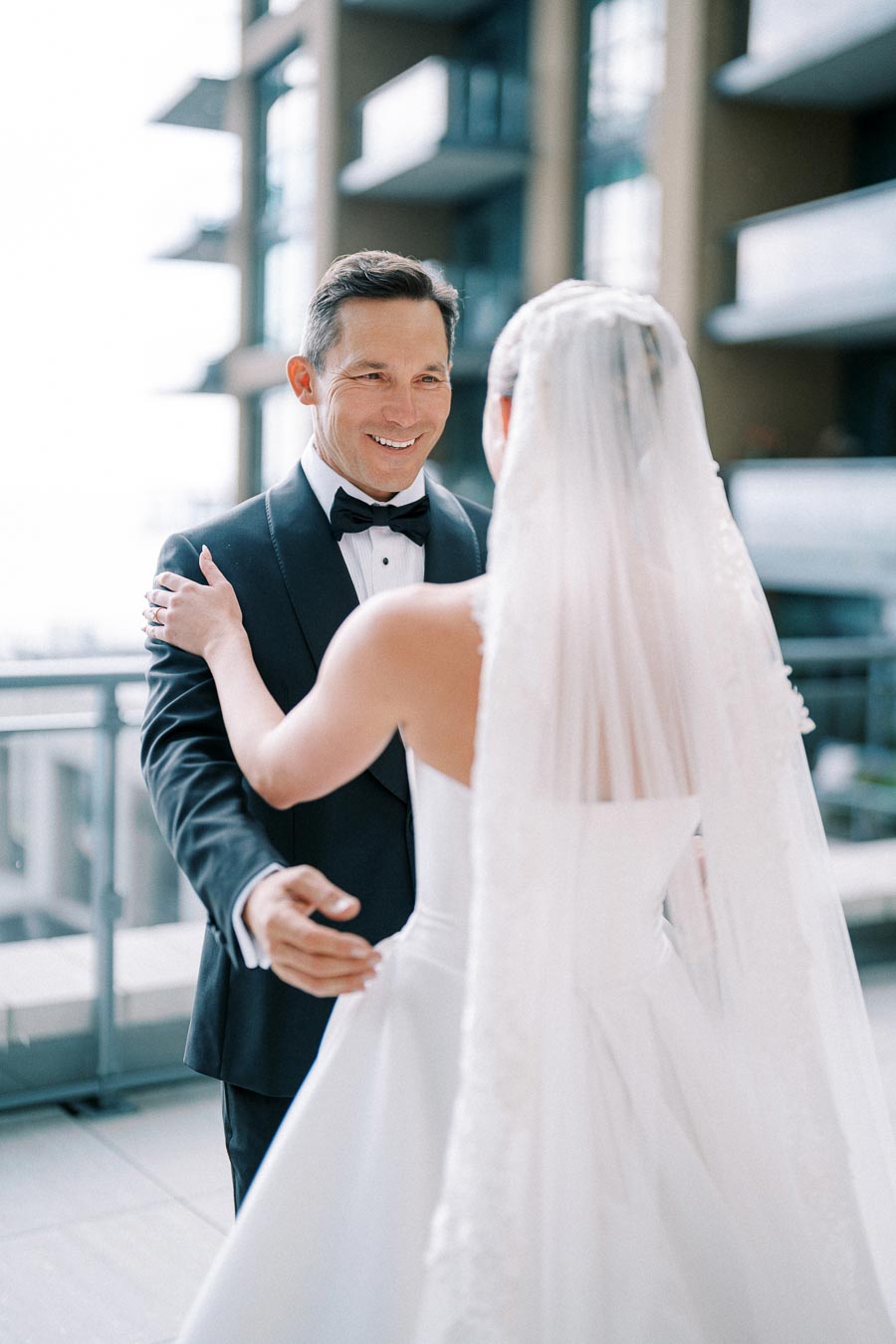A groom in a classic black tuxedo smiling at the bride during their wedding day, with a modern building backdrop.