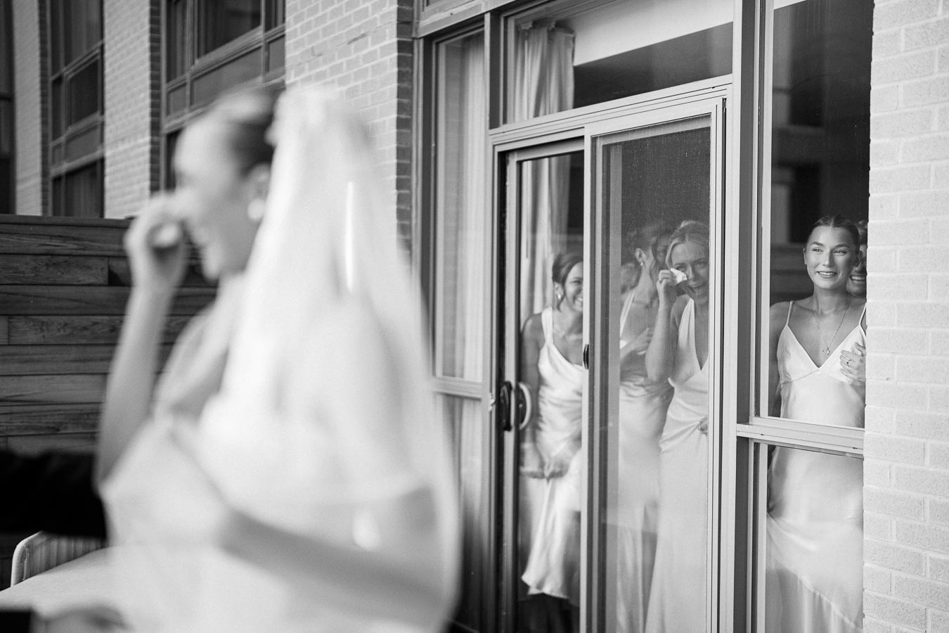 Black and white photo of a bride smiling as her bridesmaids watch through a glass door and react emotionally to her appearance.