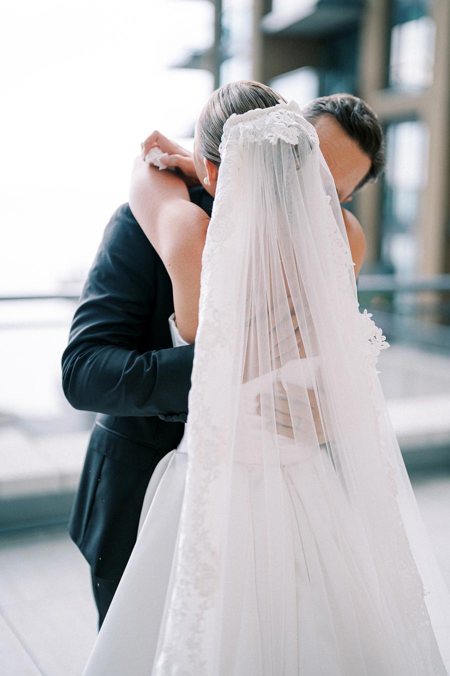 A bride and groom embrace warmly, the bride wearing a white gown and veil, during their wedding day at a modern venue with large windows in the background.