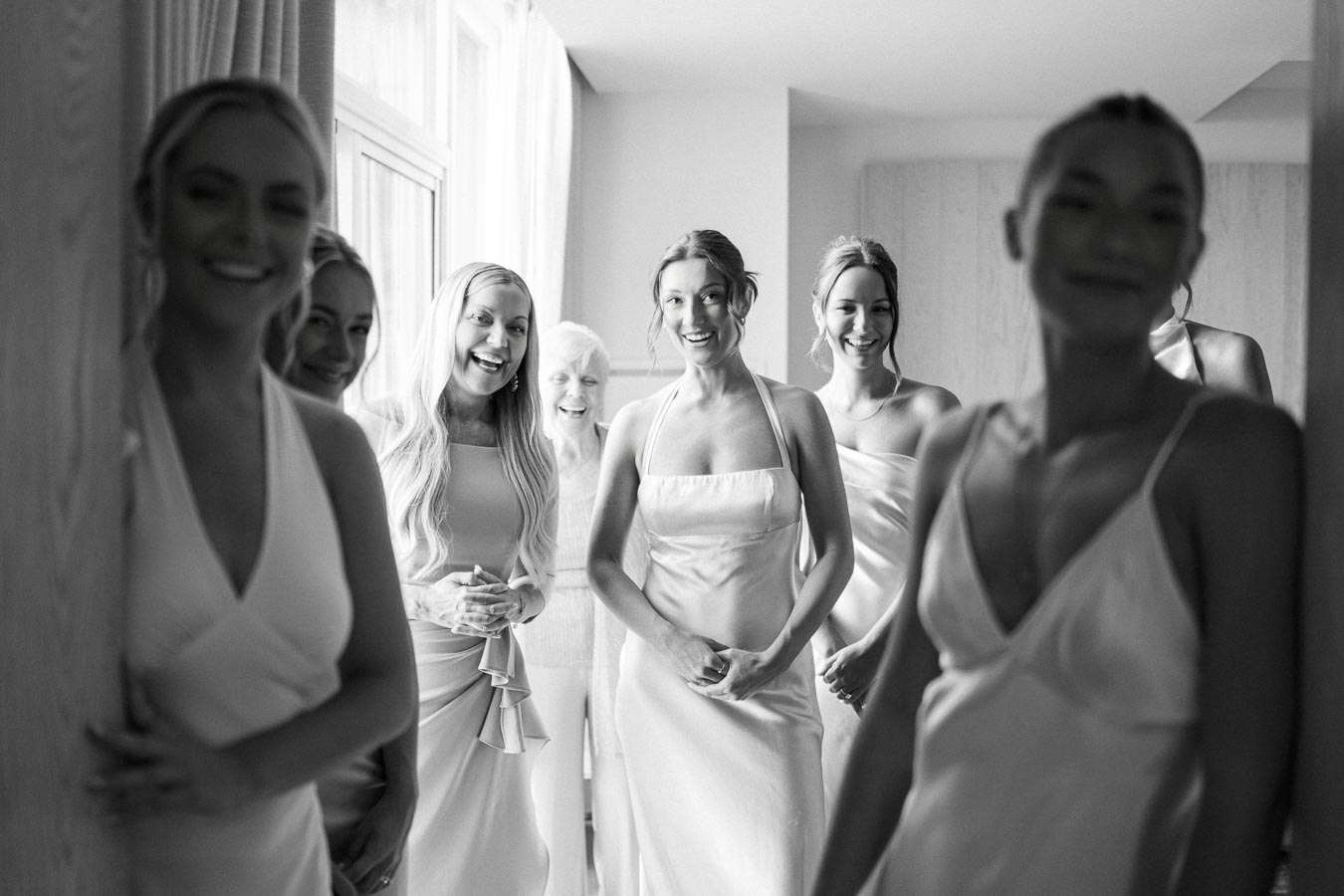 Bride surrounded by smiling bridesmaids in elegant dresses, captured in a bright, intimate setting, black and white photograph.