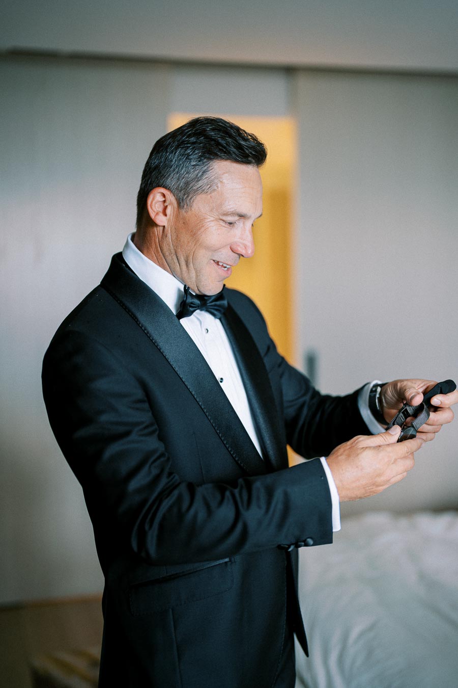 A well-dressed man wearing a classic black tuxedo and bow tie, adjusting a watch in a modern, softly lit room.