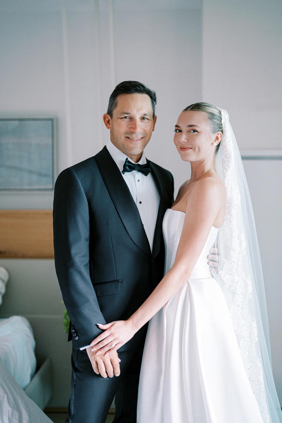 A smiling bride and groom in elegant wedding attire, with the bride in a white dress and veil, standing in a bright room.