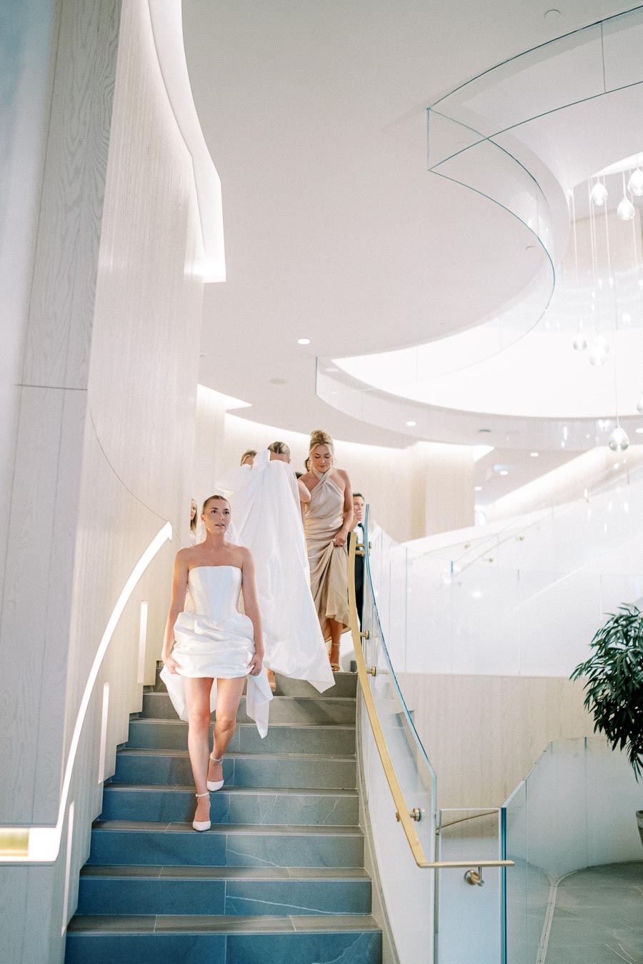 Bridal party descending a modern staircase in an elegant wedding venue, featuring a bride in a stylish white dress and bridesmaids in champagne gowns.