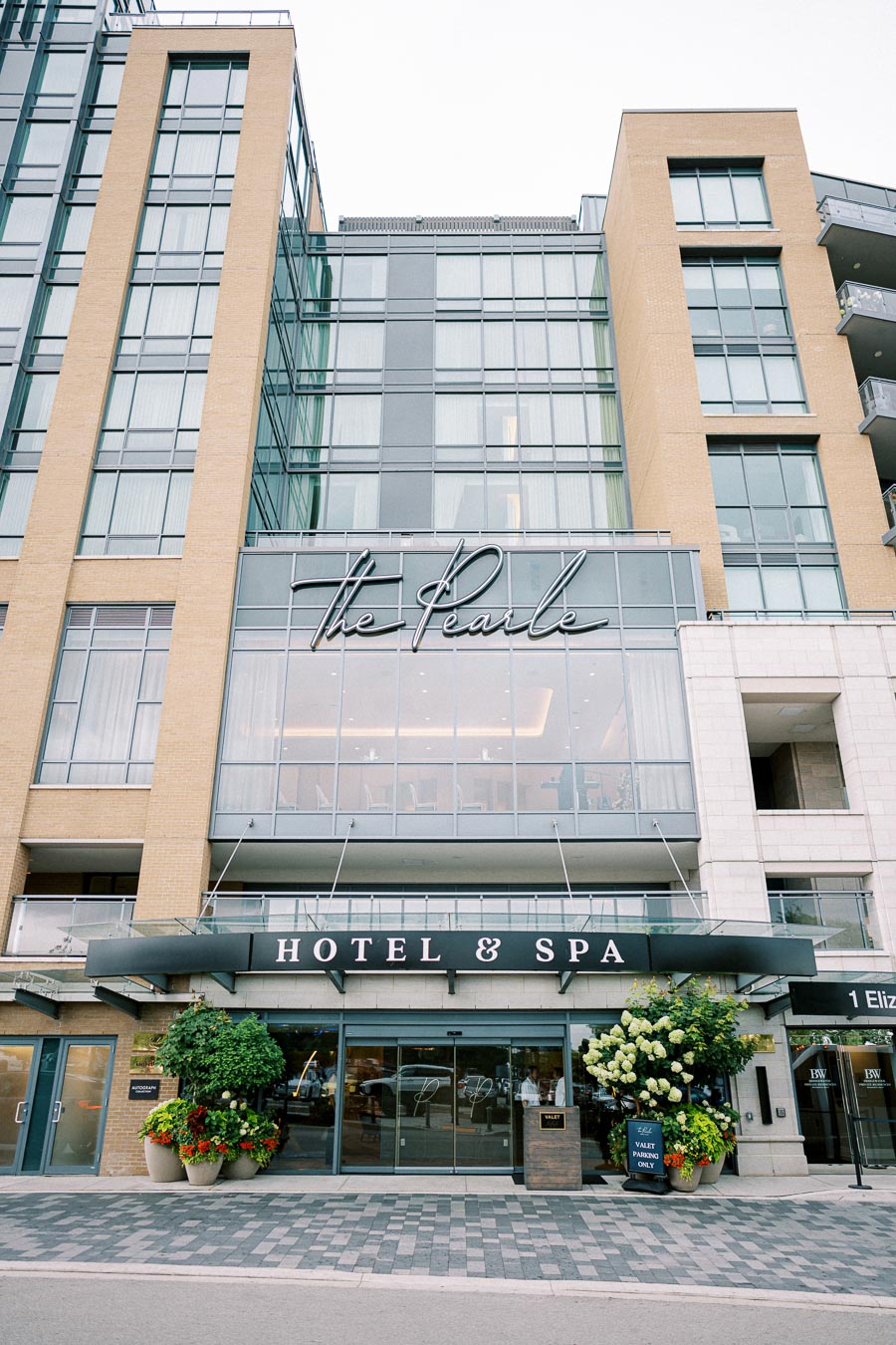 Front entrance of The Pearle Hotel and Spa, showcasing modern architecture with large windows, sleek signage, and potted plants enhancing the welcoming facade.