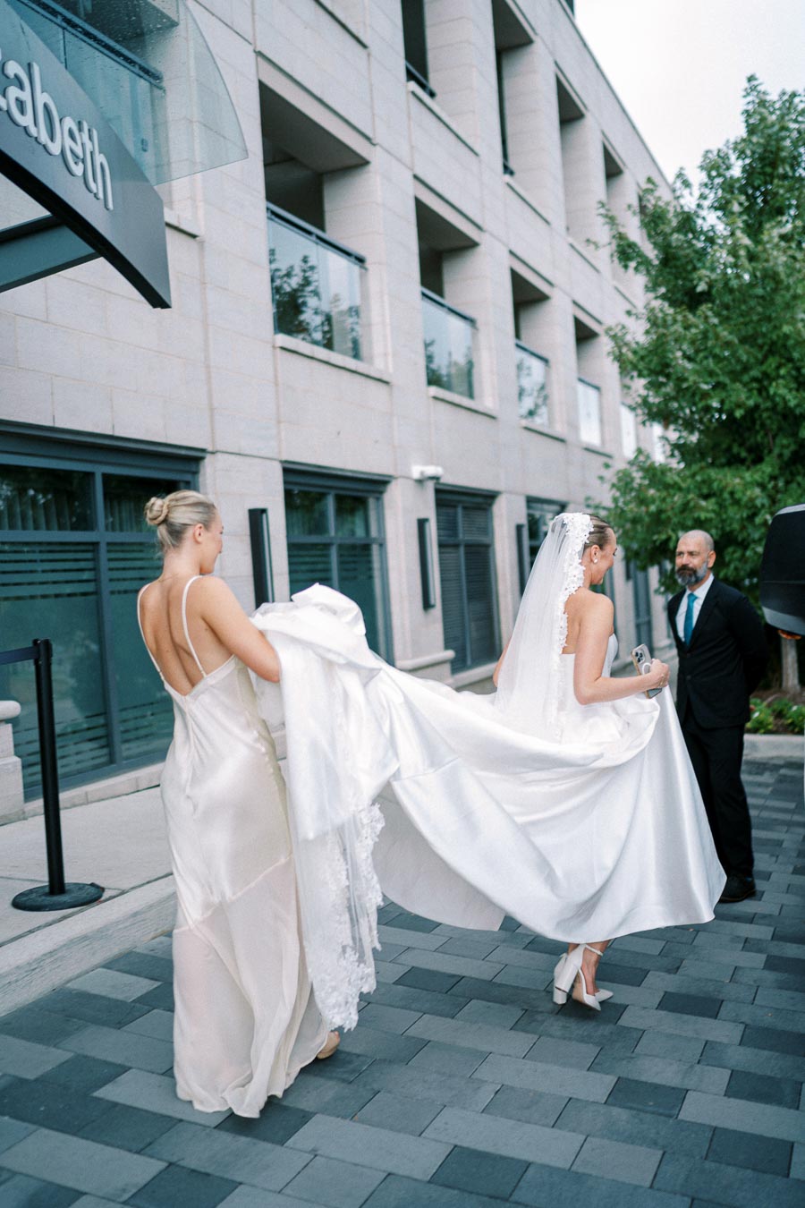 A bride in a white wedding gown walks outside a modern building, assisted by a bridesmaid in a silk dress, with a bearded man dressed in a suit nearby.