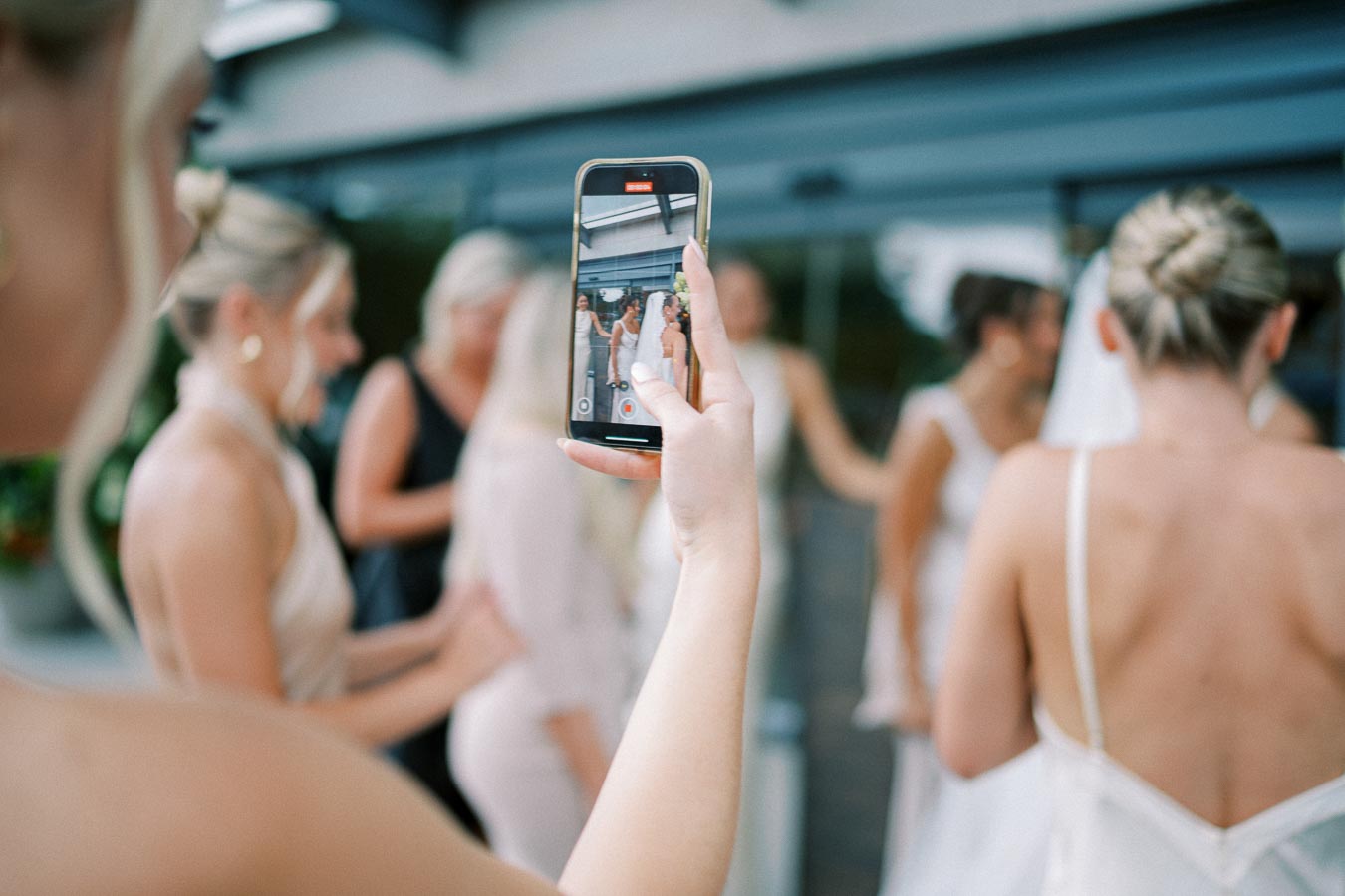 A person captures a wedding moment on their smartphone, focusing on a bride surrounded by bridesmaids in elegant dresses during an outdoor celebration.