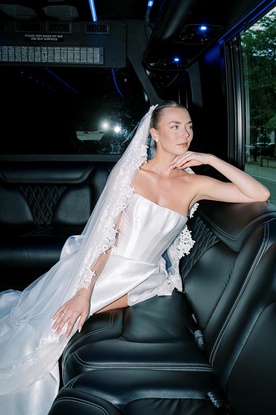 Bride in elegant satin wedding dress with lace veil, sitting in a luxury limo, gazing thoughtfully out the window.